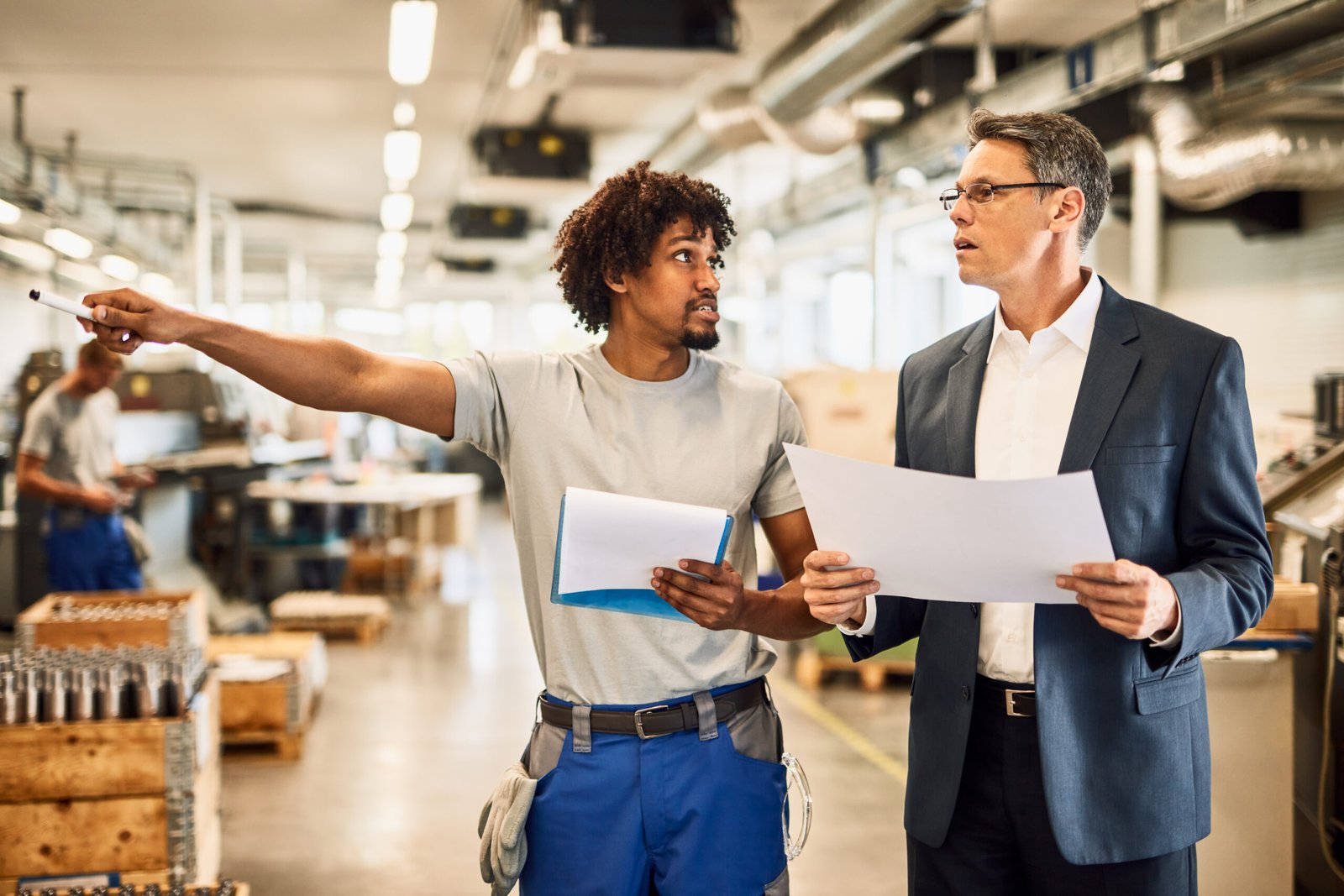 African American worker talking with company manager while leading him through the factory. Mid adult manager and black worker analyzing project plans while walking through industrial facility.